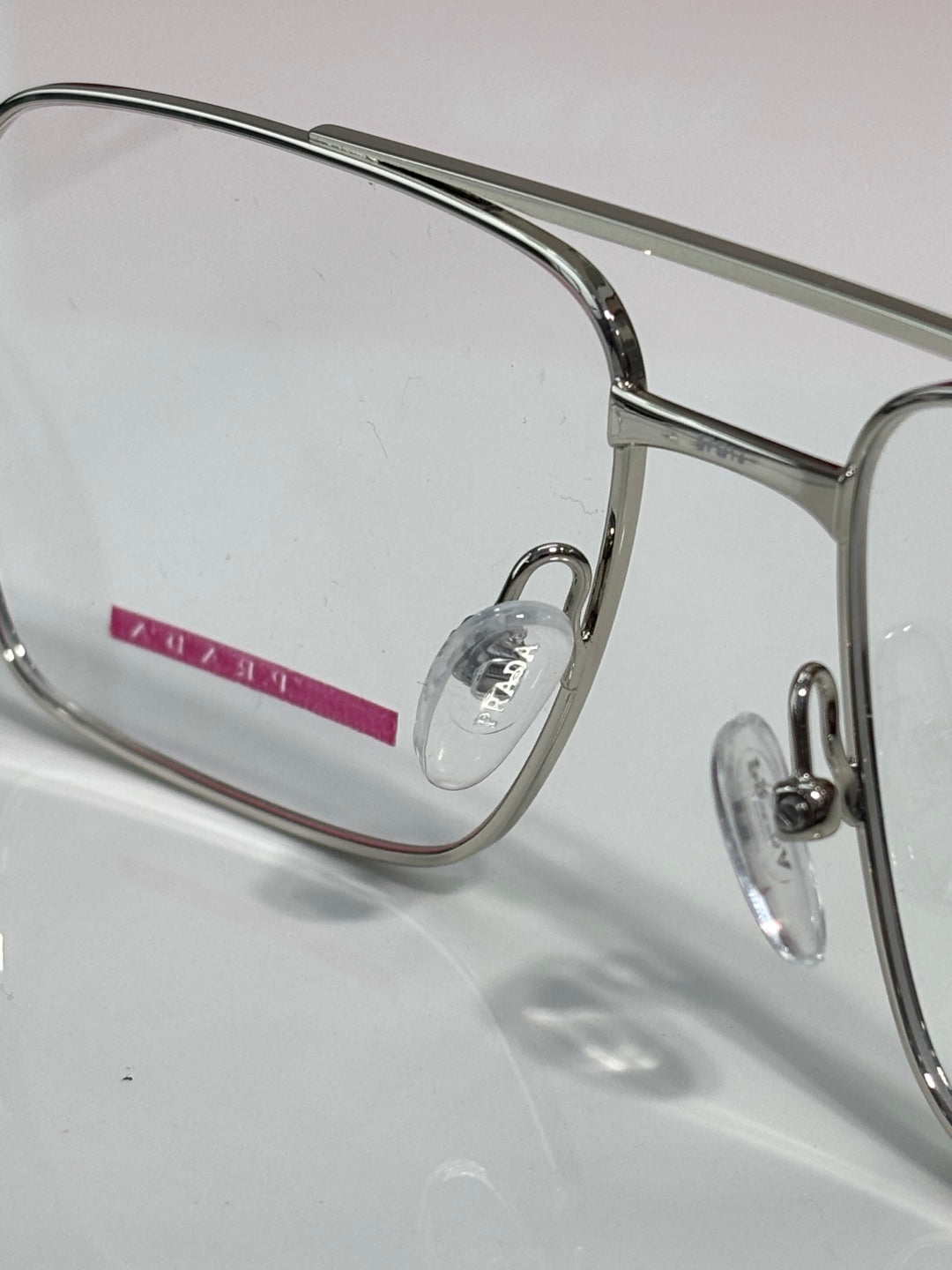 Prada eyeglasses close-up: clear lens and metal frame with small red logo detail.