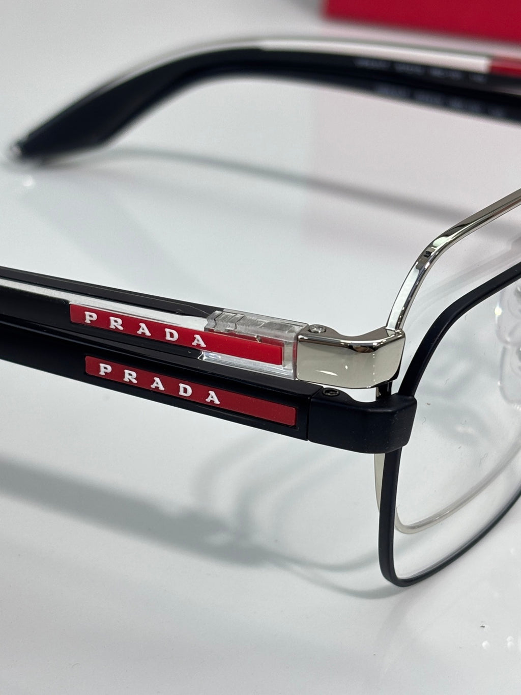 Prada eyeglasses close-up: black top bar with red “PRADA” lettering on the frame.