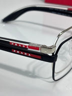 Prada eyeglasses close-up: black top bar with red “PRADA” lettering on the frame.