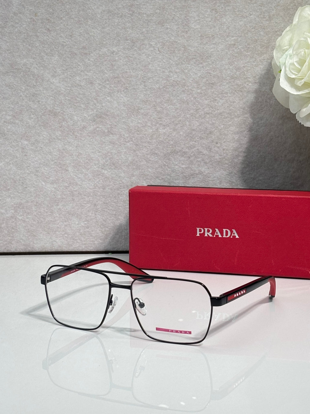 Prada eyeglasses with clear lenses and black rectangular frame, red temple logo accents.