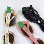 Assorted sunglasses with green floral charms, including cream, pink, and black frames