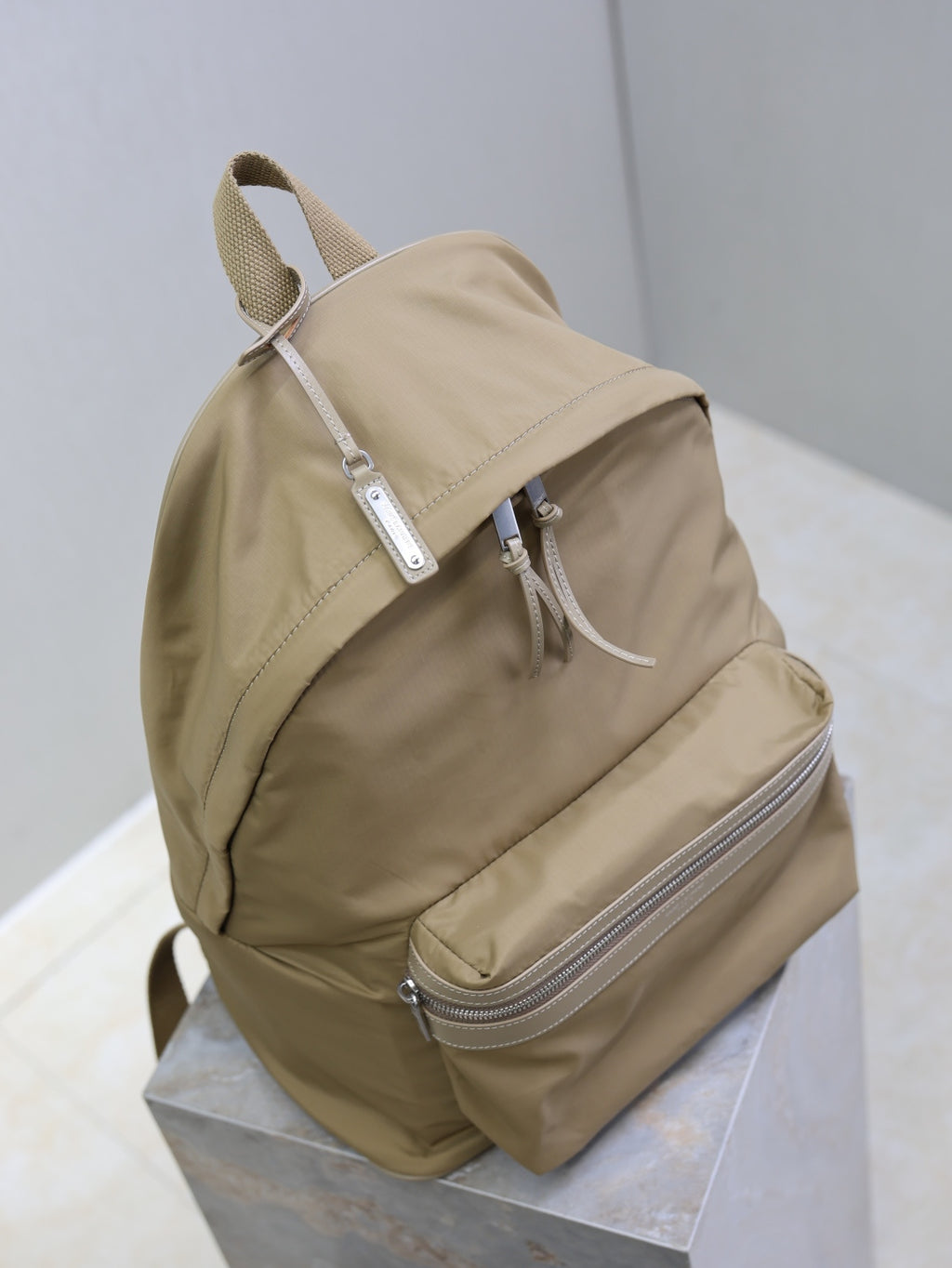 Tan nylon backpack with front-facing straps, top handle loop, and silver zip front pocket