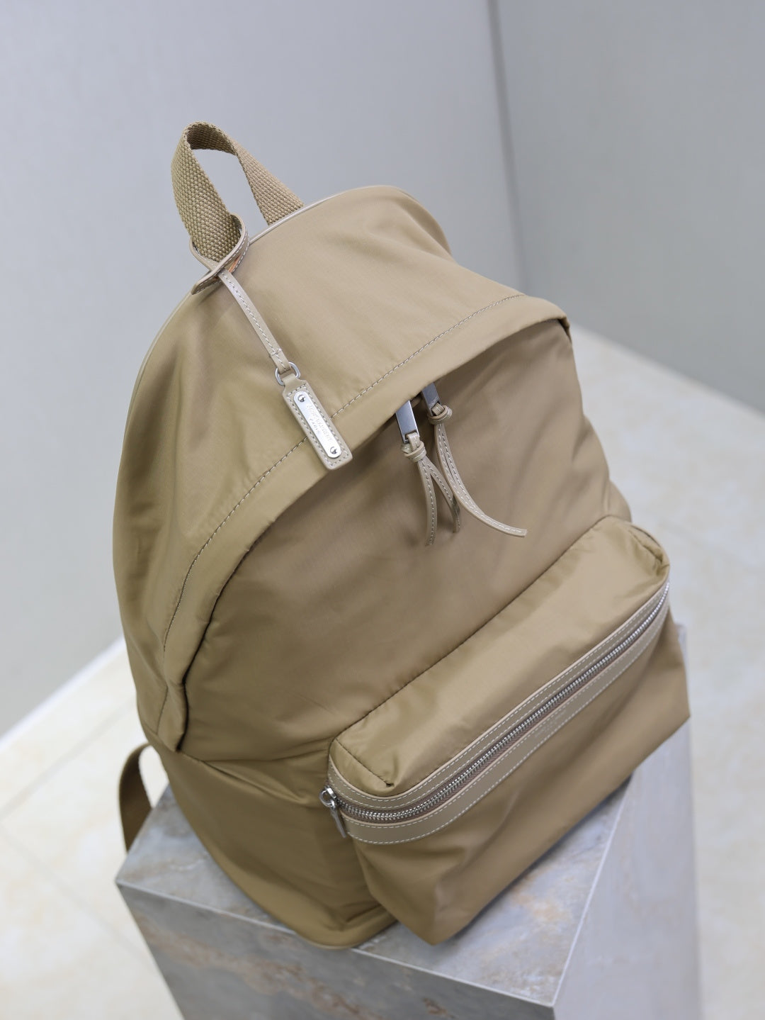Tan nylon backpack with front-facing straps, top handle loop, and silver zip front pocket