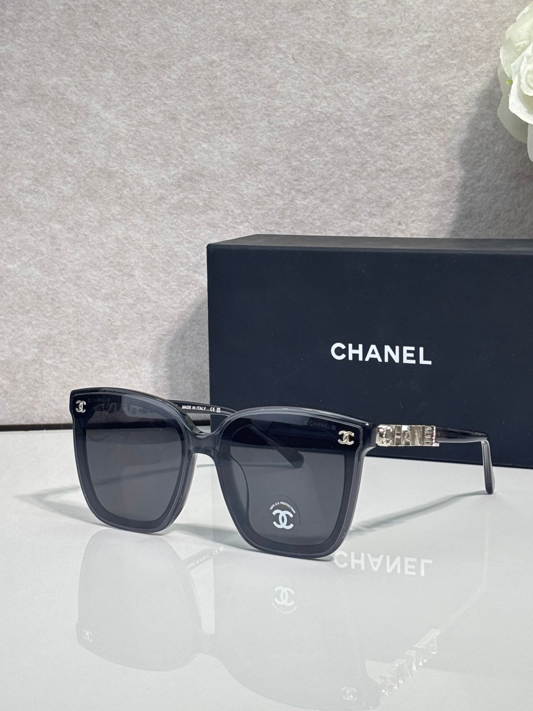 Square sunglasses with black frames and dark gray lenses, gold logo on temple arms