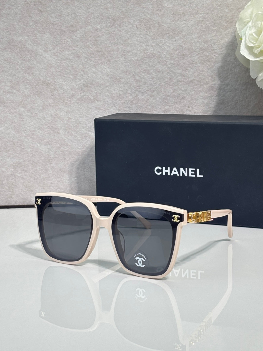 Square sunglasses with light cream frames and dark gray lenses, gold side logo detail