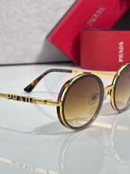 Prada round sunglasses close-up with gold-tone frame and amber-tinted lenses, tortoise tips