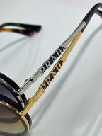 Prada sunglasses temple close-up with gold-tone bar and Prada lettering, dark lens