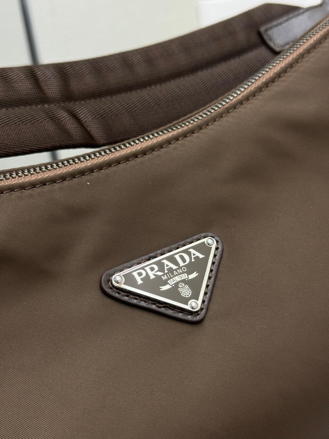 Triangular metal logo patch on brown nylon hobo bag with PRADA text