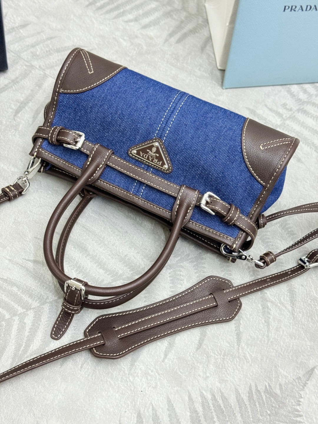 Blue denim Prada handbag laid flat with brown leather strap and logo tag