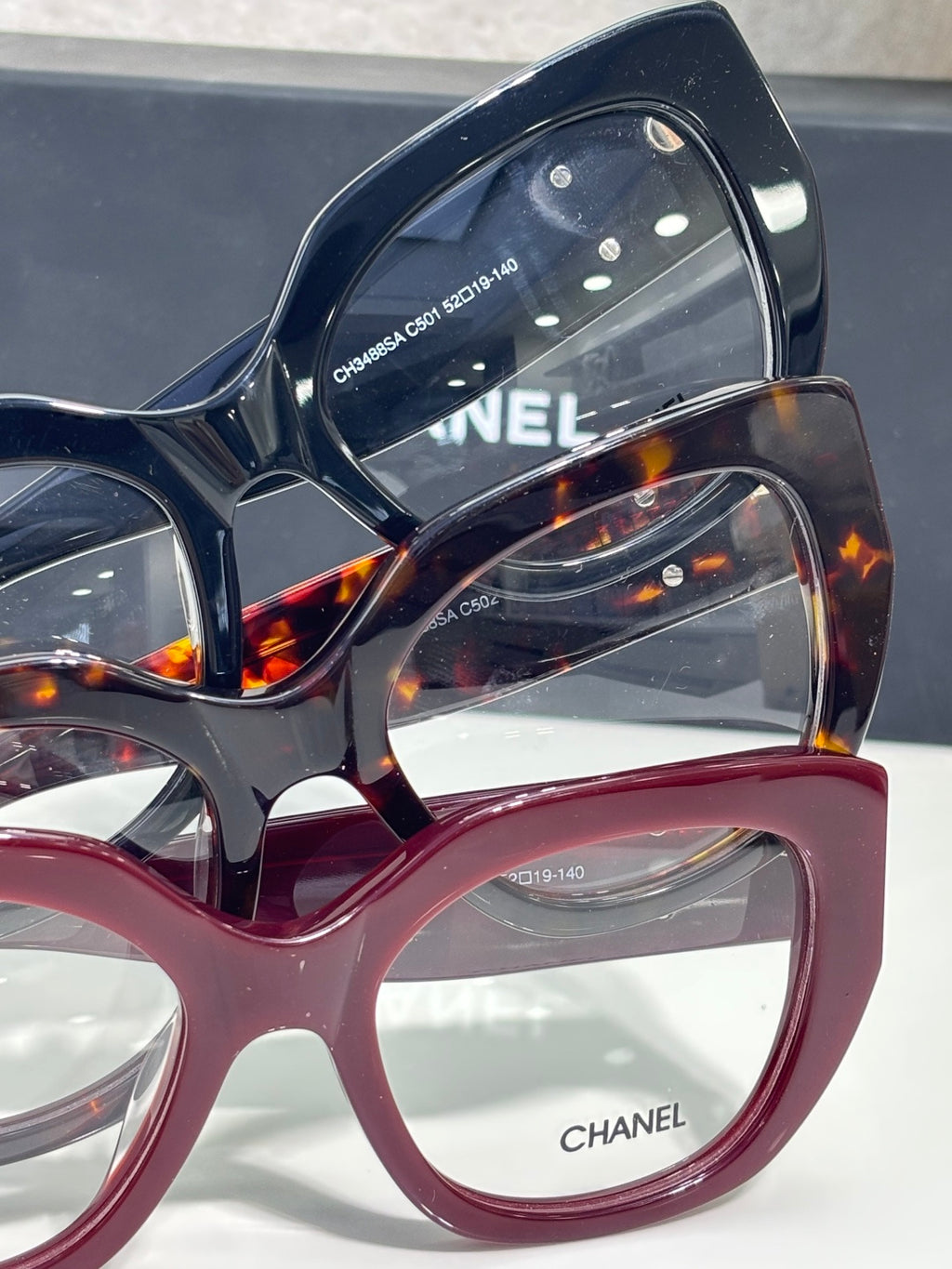 Eyeglasses close-up with dark blue frames and burgundy lower rims; gold Chanel branding.