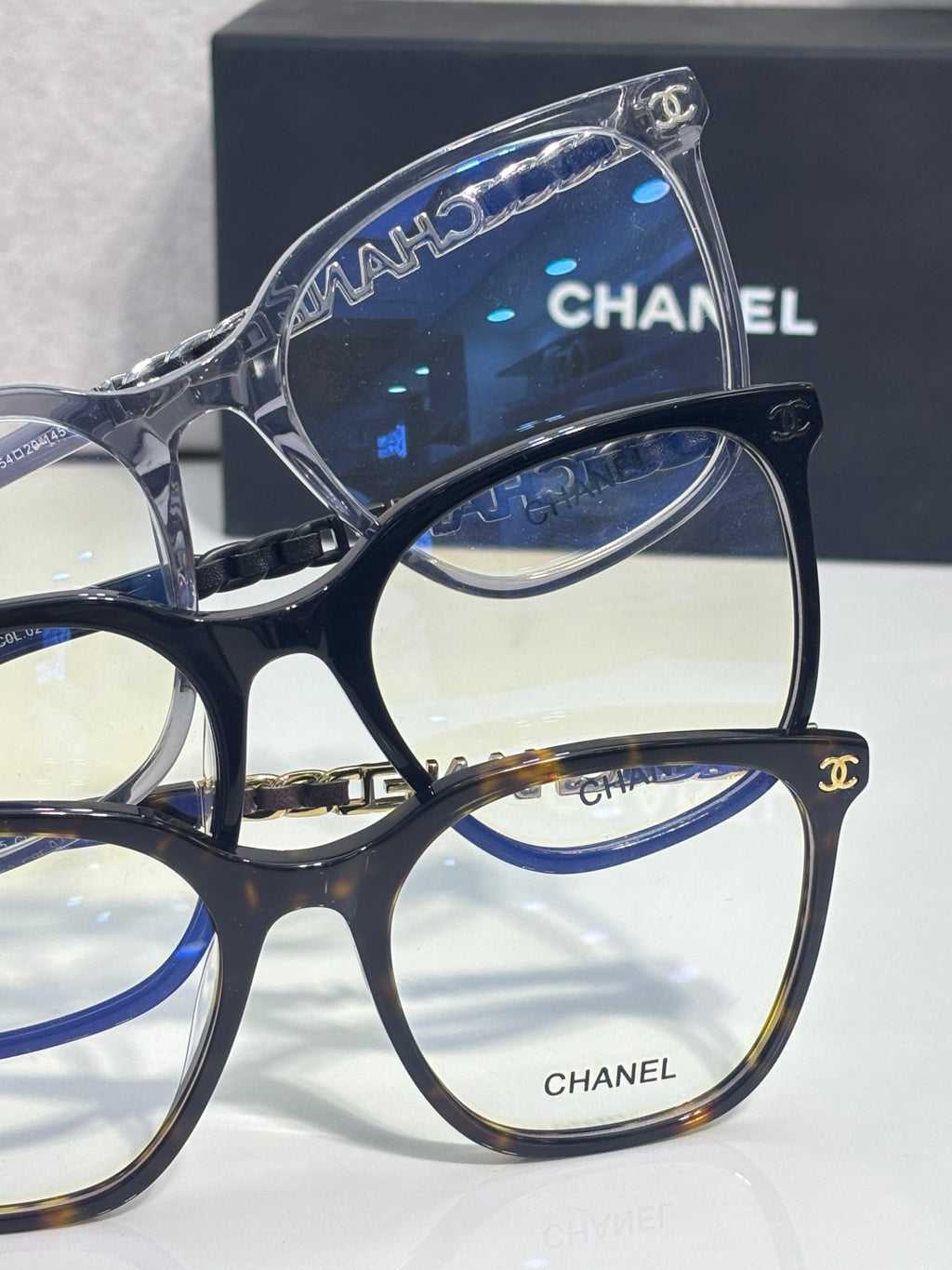 Eyeglasses close-up showing blue-tinted lens and gold CHANEL logo on frame front.