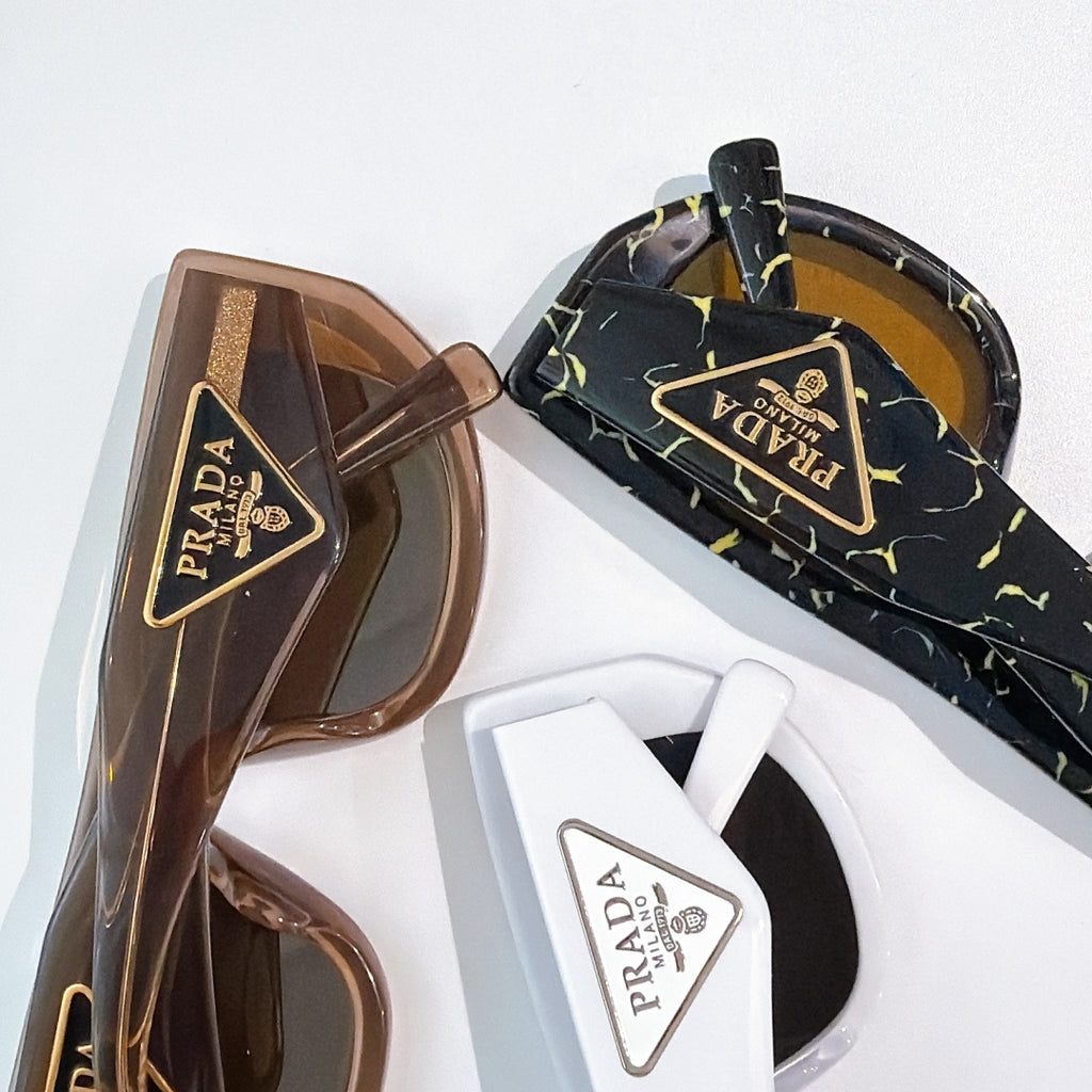 Prada sunglasses close-up: brown frames with tinted lenses and gold triangular logo detail.