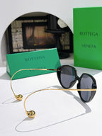 Bottega Veneta oval sunglasses with black frame and dark blue lenses, gold chain strap.