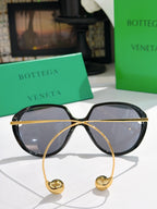Bottega Veneta sunglasses with black frame, dark lenses, and gold strap charms.