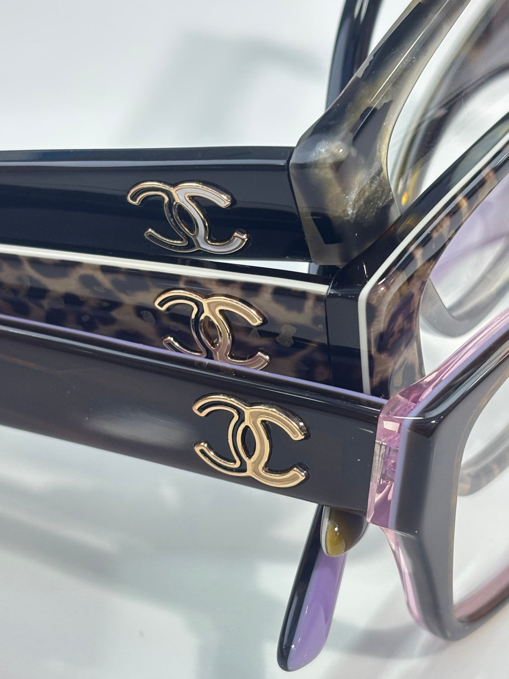 Dark eyeglasses close-up with gold CC emblems on frame and purple detail at edge.
