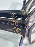 Dark eyeglasses close-up with gold CC emblems on frame and purple detail at edge.