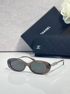 Chanel glasses with dark brown oval frames, dark lenses, and gold logo temple accents.