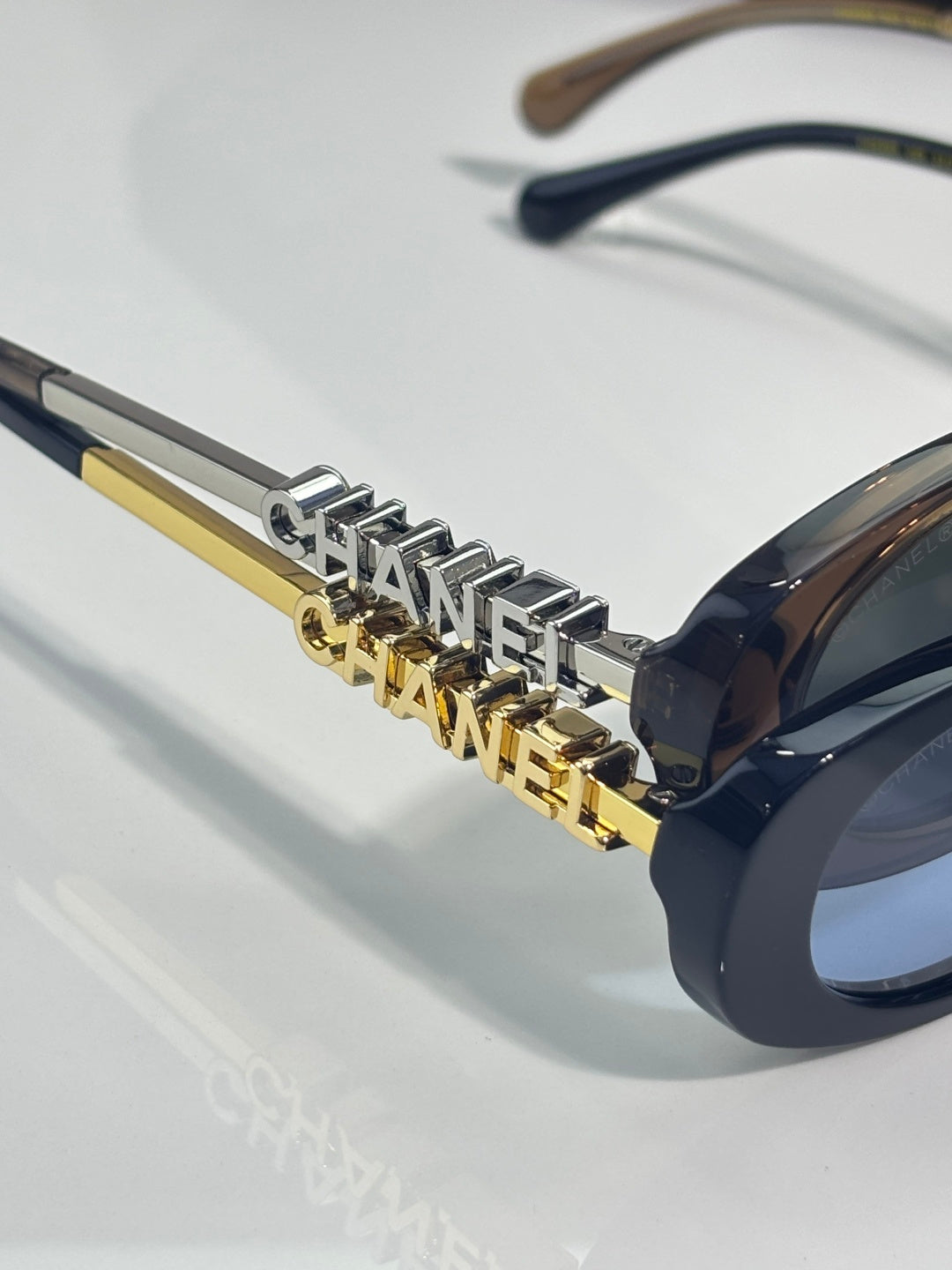 Chanel glasses close-up: gold logo link detail on dark temples near oval lens edge.