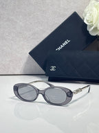 Chanel glasses with charcoal oval frames, dark lenses, and gold logo temple detailing.