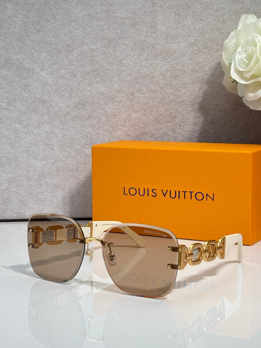 Sunglasses with neutral tinted lenses and gold frame; chain-link temple with logo detail.