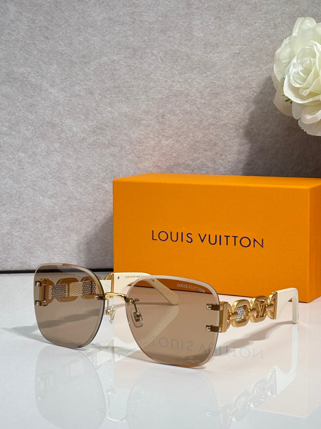 Sunglasses with neutral tinted lenses and gold frame; chain-link temple with logo detail.