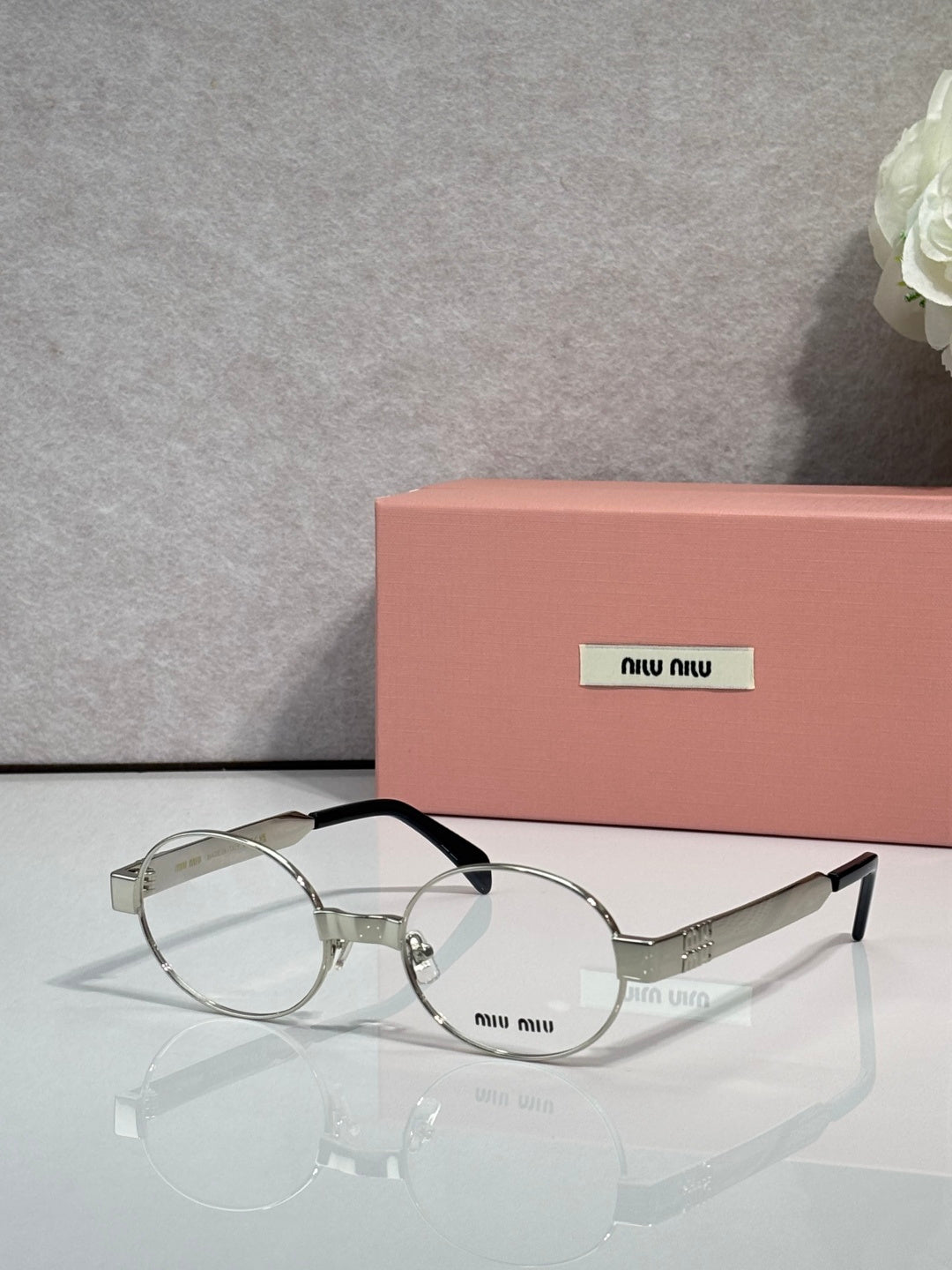 Round eyeglass frames with clear lenses, silver-toned frame and black temples, Miu Miu logo.