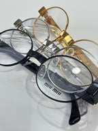 Close view of eyeglass frames with clear lenses, gold and black temples, Miu Miu logos.