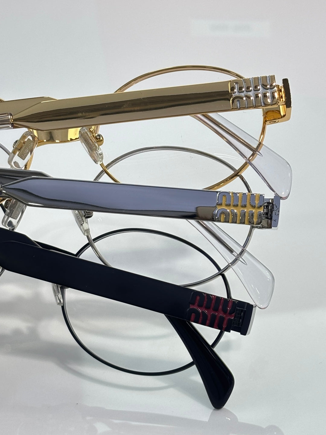 Close view of multiple eyeglass frames: gold arms and dark blue temples, Miu Miu logos.