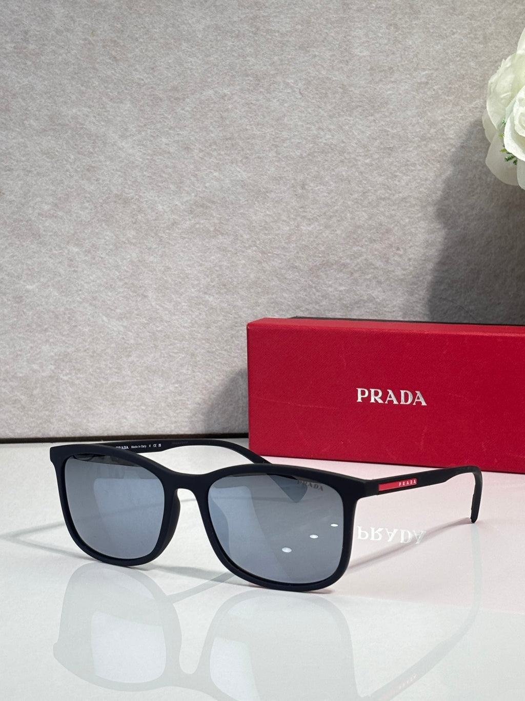 Prada sunglasses with dark lenses and black frames, beside red Prada-logo case