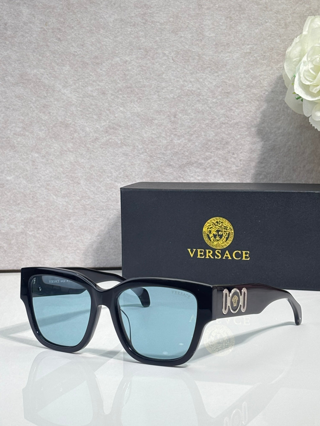 Versace sunglasses with dark frame and light blue lenses, gold “101” emblem on temple.