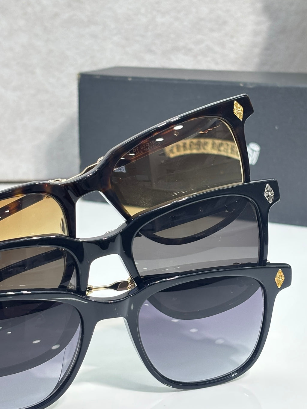 Sunglasses close-up with dark frames, gradient lenses, and gold temple emblems.