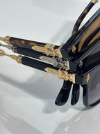 Sunglasses close-up: dark tortoise-style frame with gold ornate temple hardware.