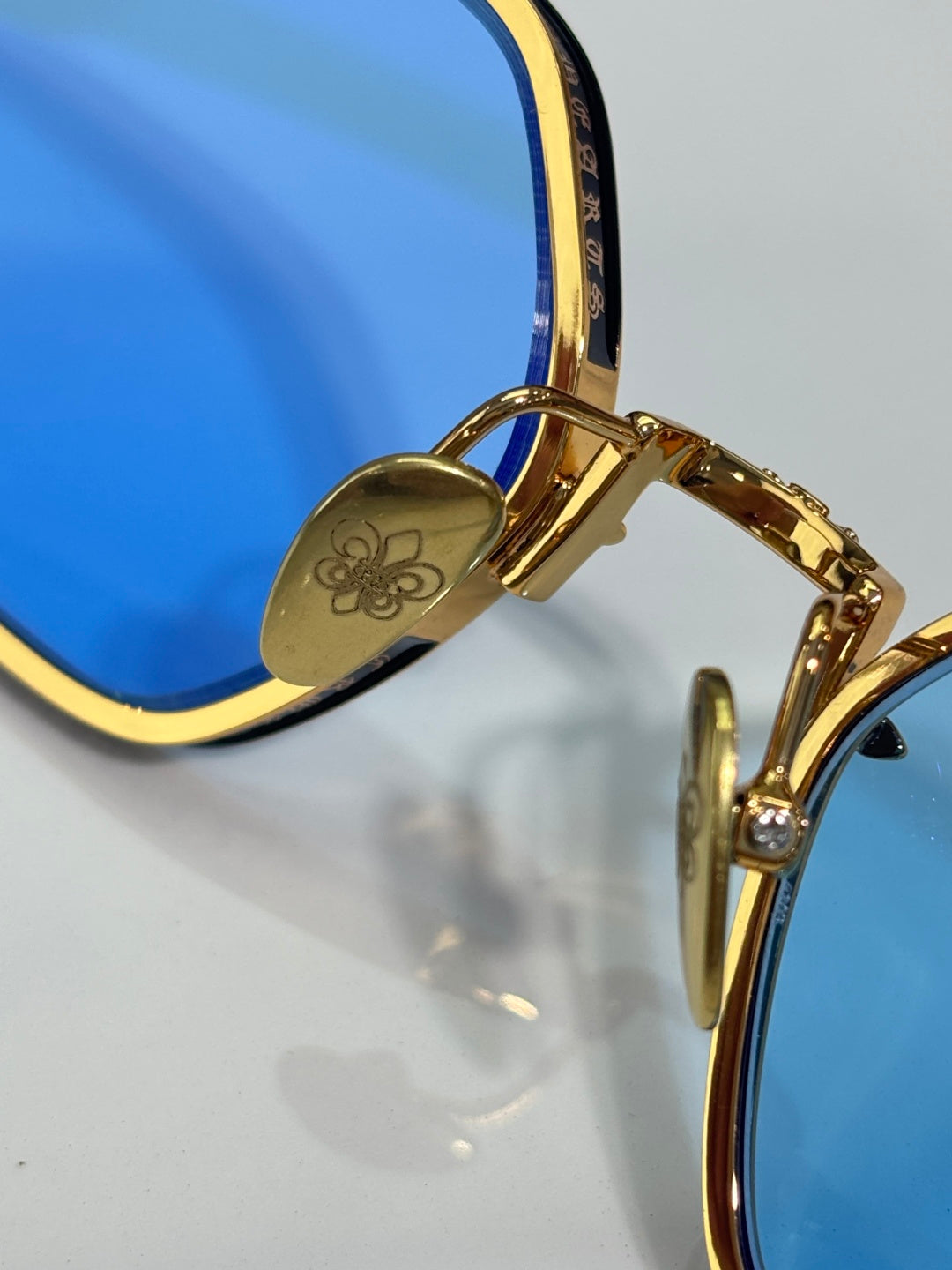 Gold-framed sunglasses hinge detail with decorative emblem and clear lens edge