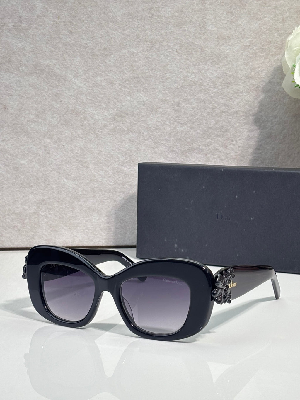 Black Dior glasses with rounded square lenses and white floral charm on the temple.