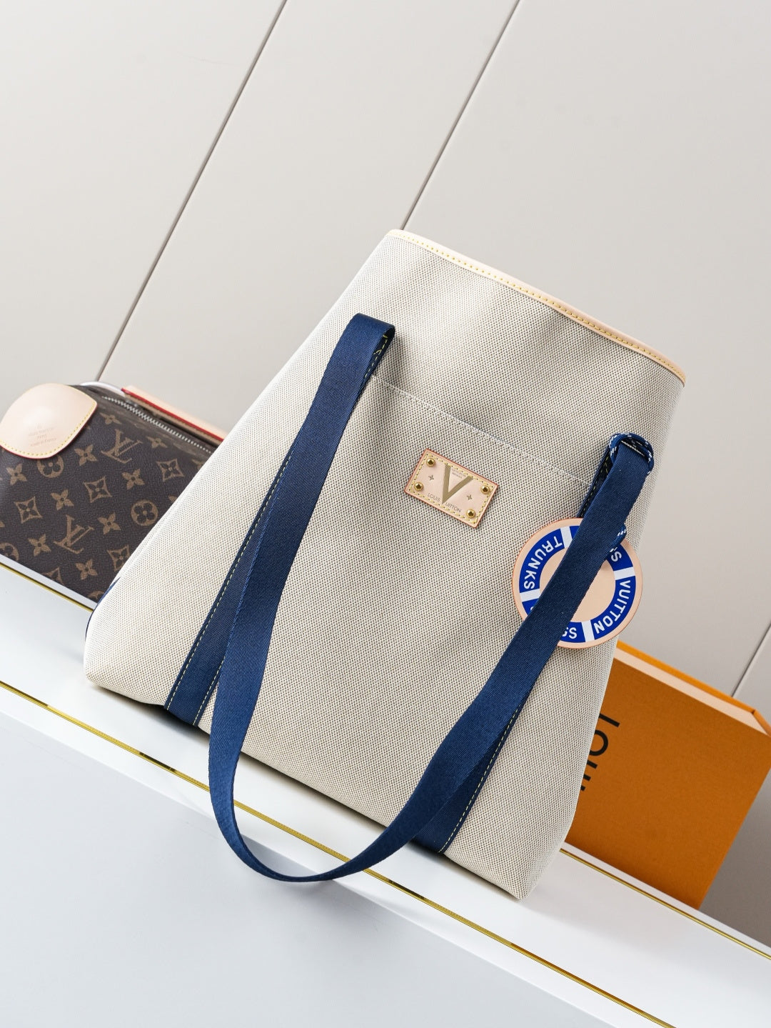 Beige canvas tote bag with navy handles and central monogram patch, gold hardware