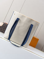 Beige canvas tote bag with navy vertical handle straps and gold corner studs