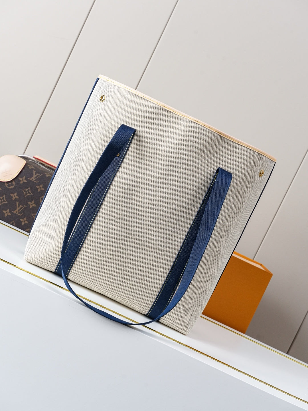 Beige canvas tote bag with navy vertical handle straps and gold corner studs