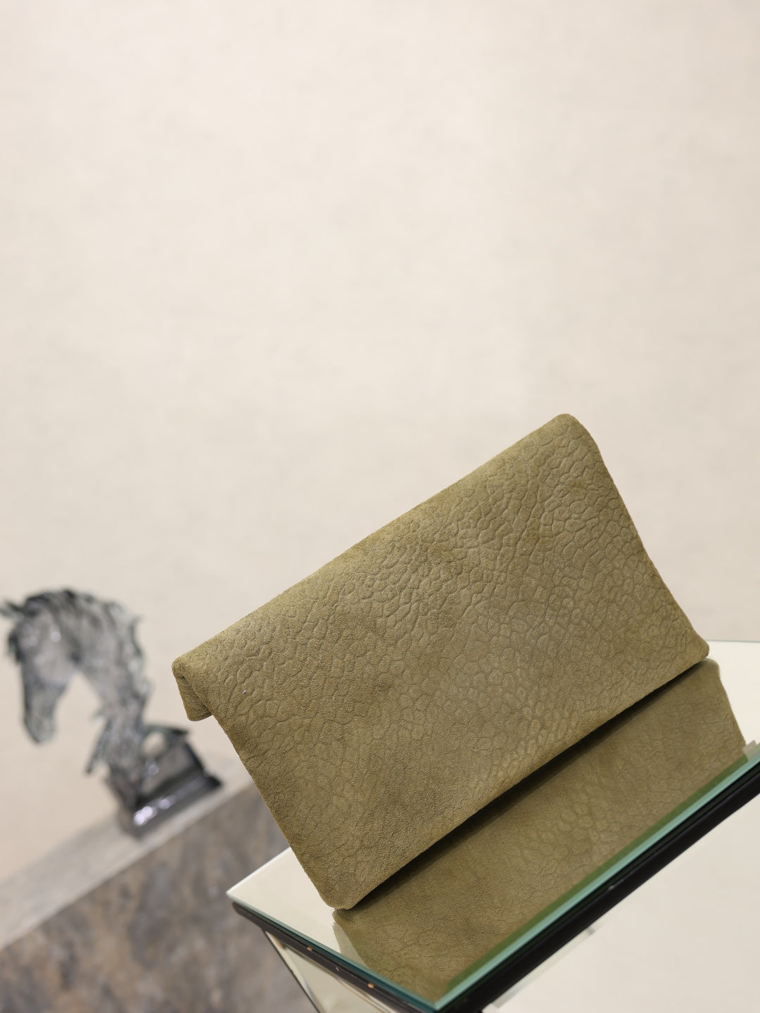 Olive textured leather envelope clutch bag resting on a glass pedestal