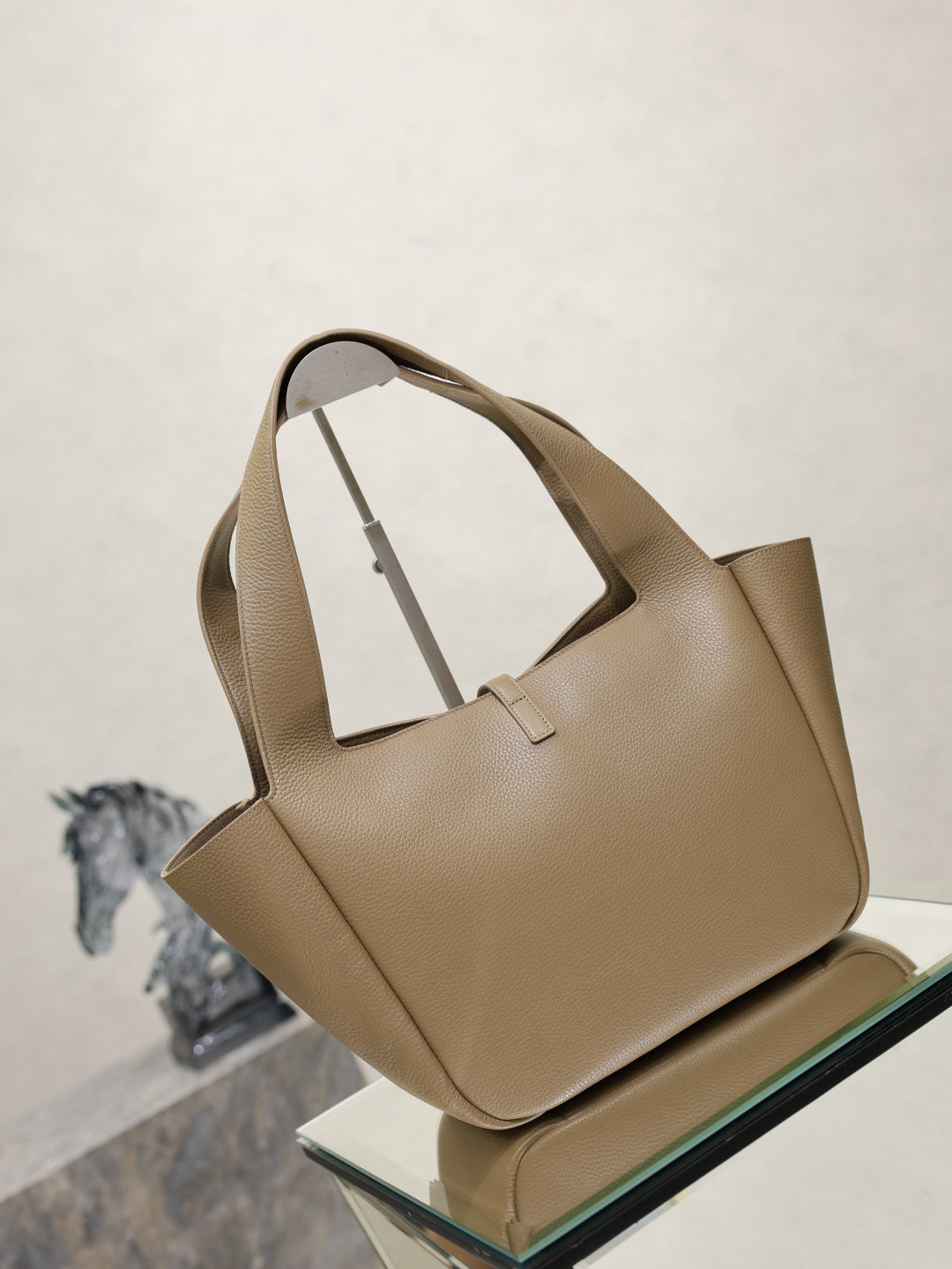 Beige leather tote bag with top handles and gold YSL logo charm, standing on glass