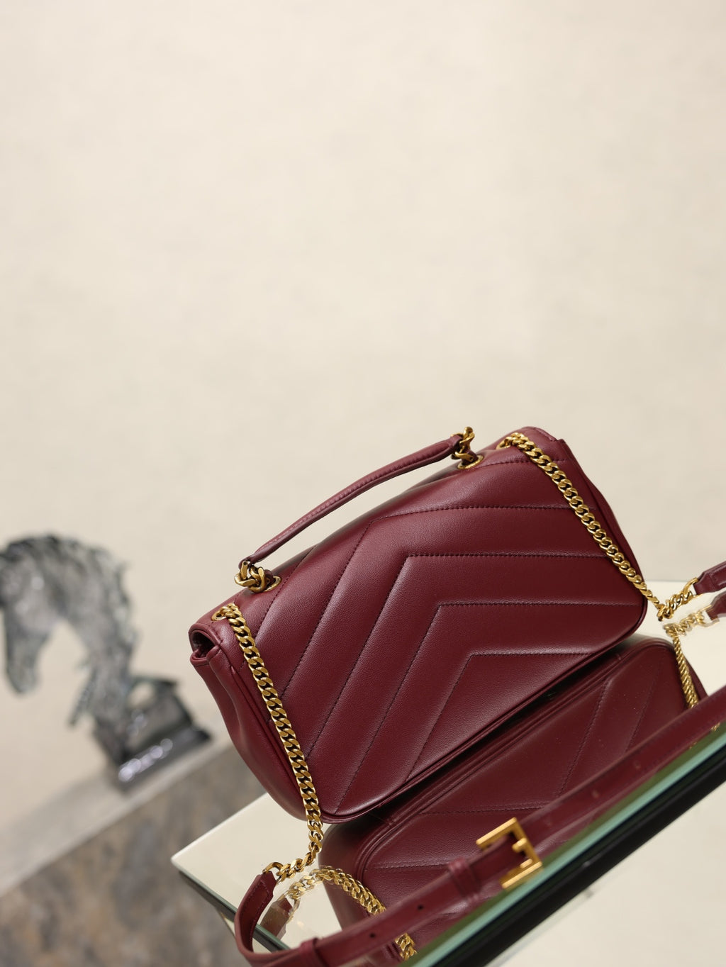 Burgundy quilted shoulder bag with front flap and gold chain strap on glass display.