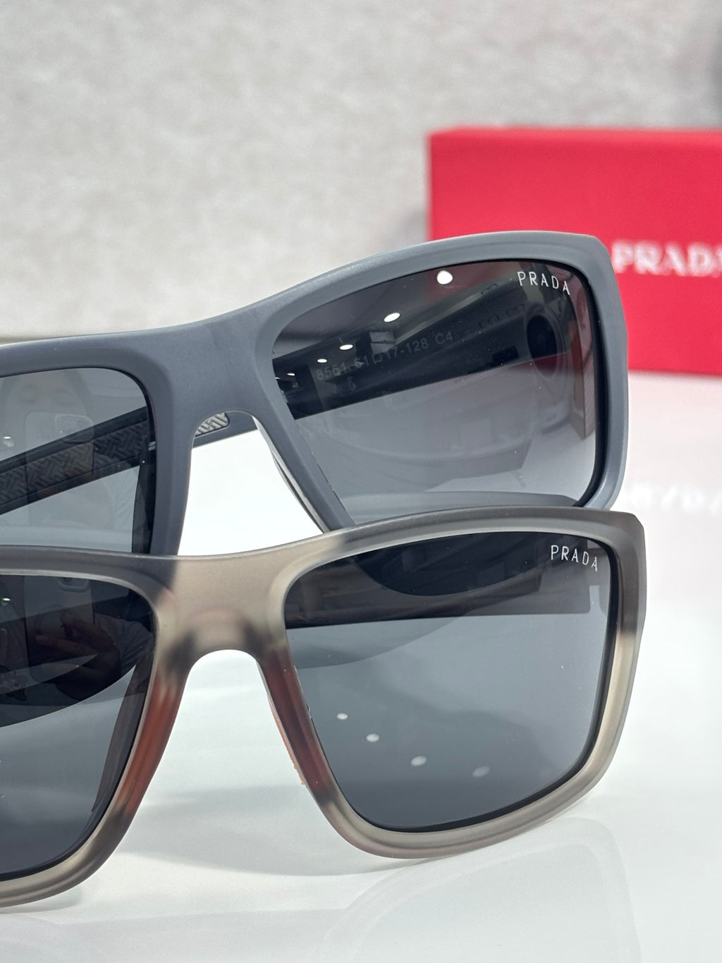 Prada sunglasses pair with light gray frames and dark lenses, with red Prada case