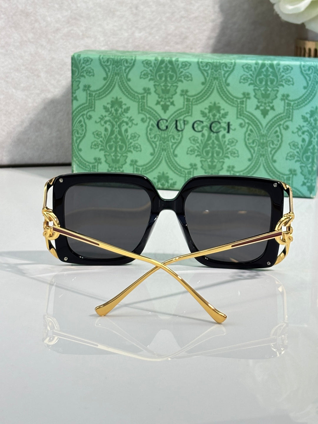 Black square sunglasses on clear stand, dark lenses with gold-tone frame details.