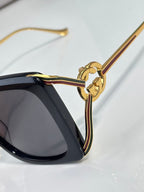 Close-up: black sunglasses with gold-tone circular logo charm and temple arm.