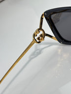 Black sunglasses with gold-tone arm and circular logo detail; dark lens.
