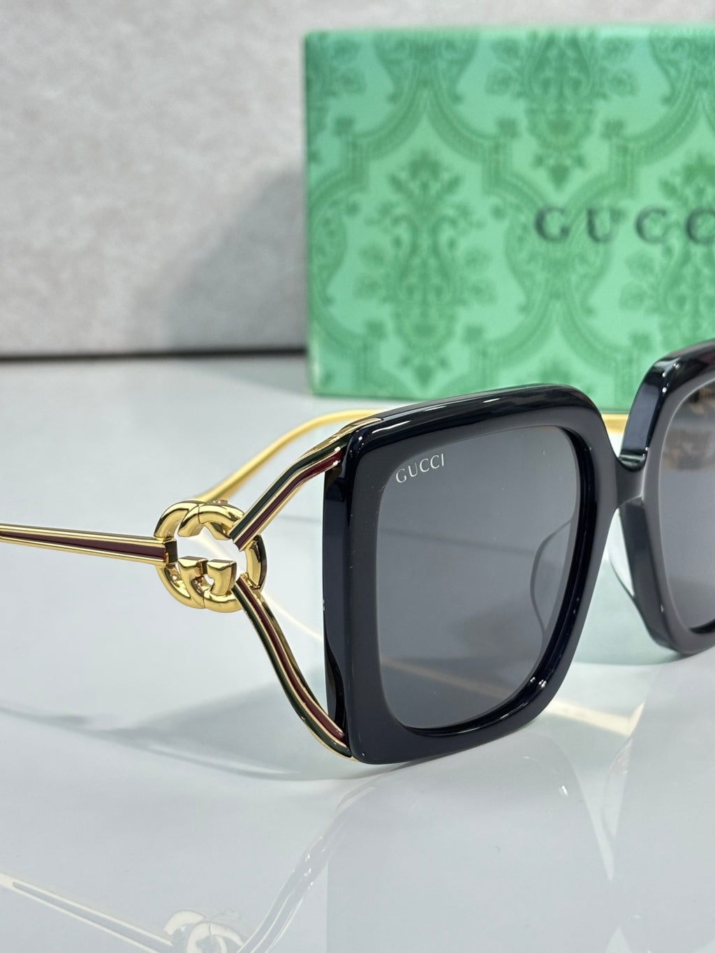 Black sunglasses with gold-tone temple arm and circular logo charm; dark lens.