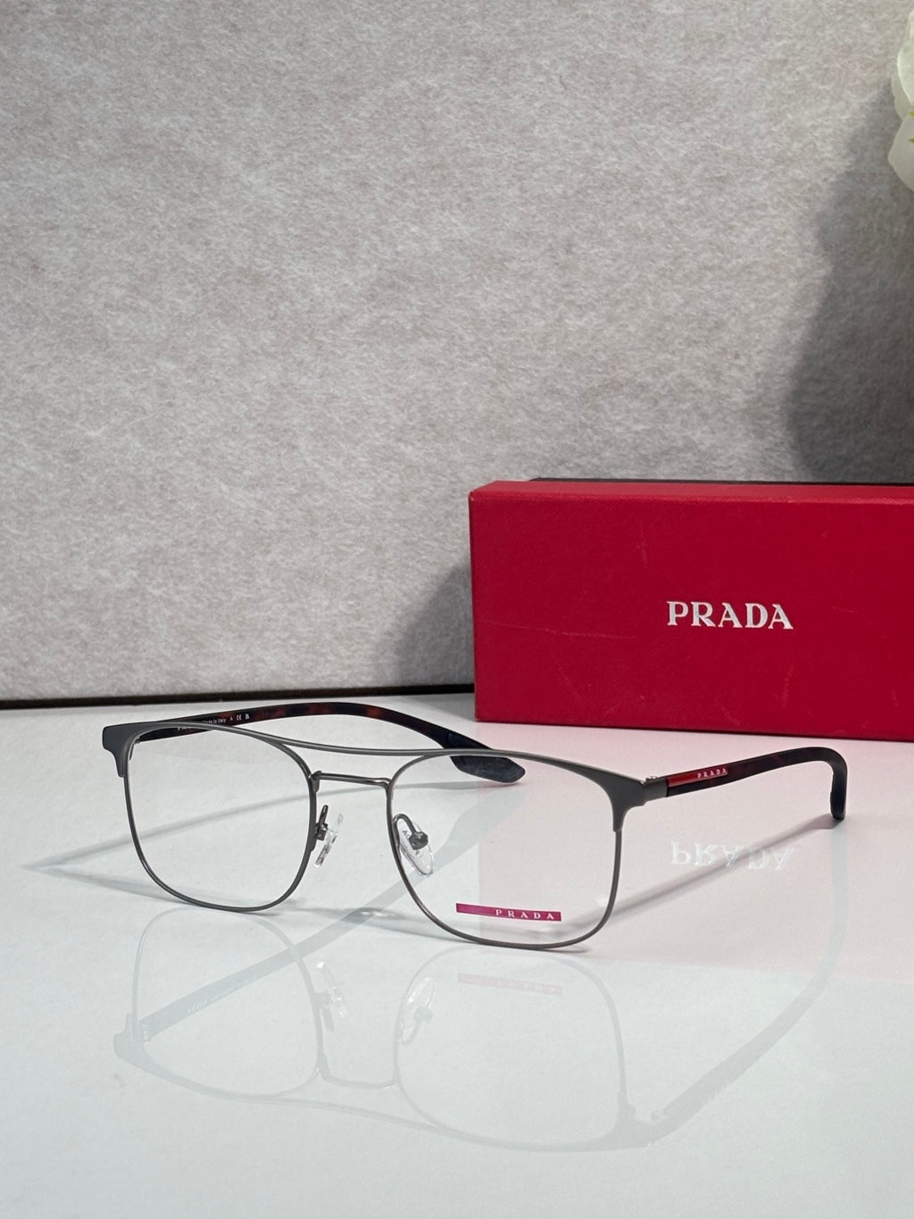 Black full-rim eyeglasses with clear lenses and red logo accents, near a Prada case.