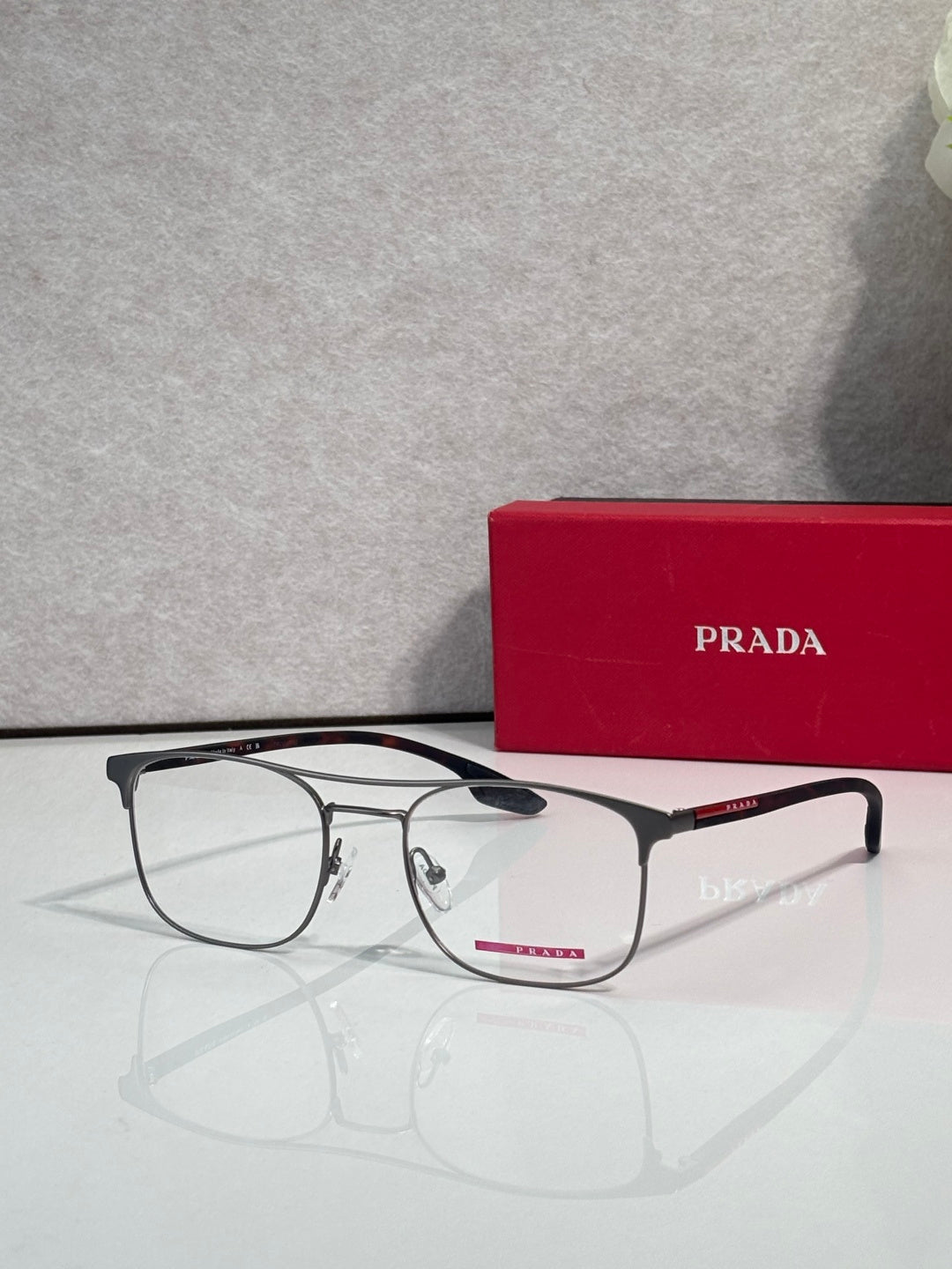 Black full-rim eyeglasses with clear lenses and red logo accents, near a Prada case.