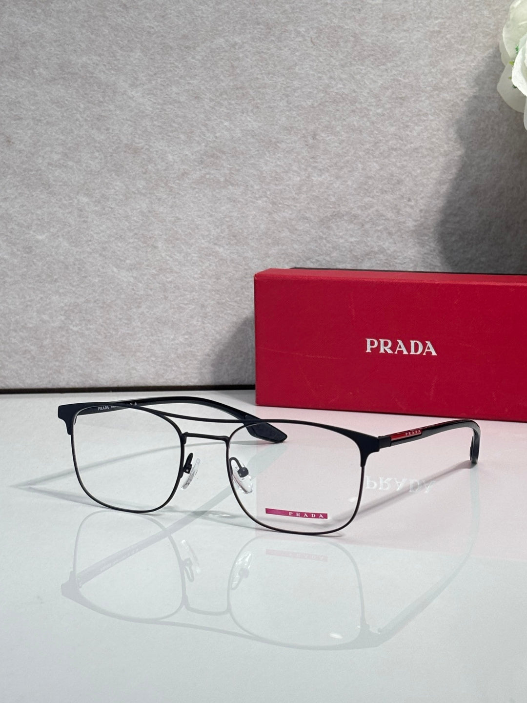 Black full-rim eyeglasses with clear lenses and red logo accents, with a red Prada case.