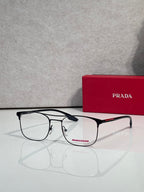 Black full-rim eyeglasses with clear lenses and red logo accents, beside a Prada case.