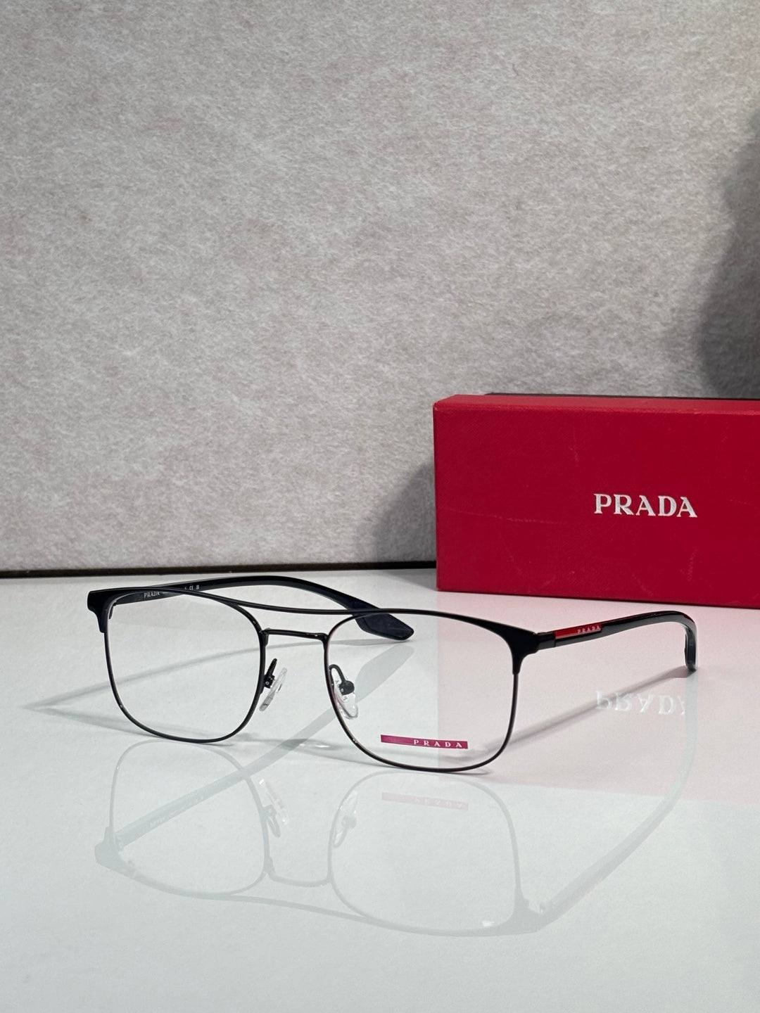 Black full-rim eyeglasses with clear lenses and red logo accents, beside a Prada case.